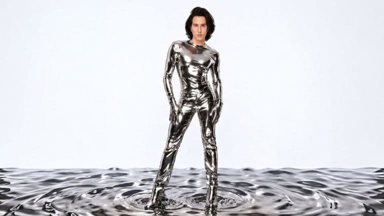 Ricky Rebel stands in a reflective chrome outfit on a metallic liquid surface against a bright backdrop.
