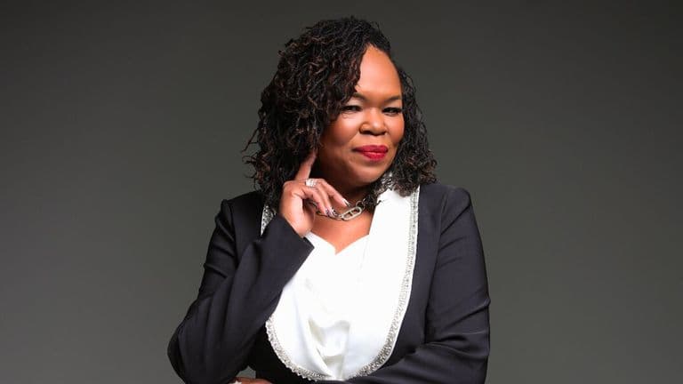 A confident woman in a black blazer with silver trim and a white blouse stands against a grey background, smiling warmly with her hand near her chin.