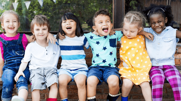 Diverse group of young children sitting together outdoors, smiling and laughing with arms around each other.