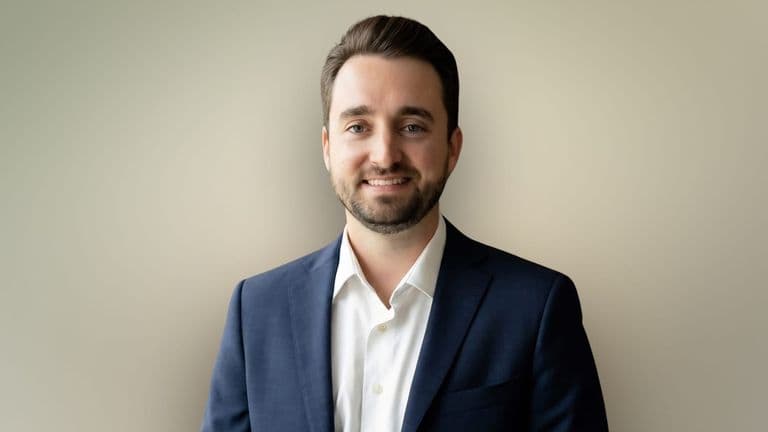 Professional headshot of smiling man in navy suit and white shirt against neutral background.