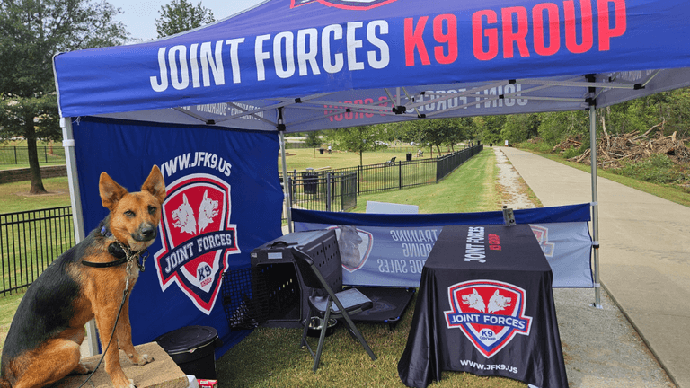 German shepherd at Joint Forces K9 Group booth under blue canopy in park setting.