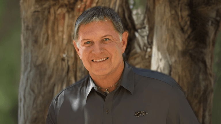 Close-up portrait of a smiling man with short hair, wearing a grey shirt, standing outdoors in front of a tree, showcasing a positive and confident expression.