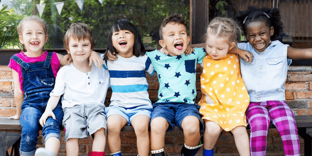 Diverse group of young children sitting together outdoors, smiling and laughing with arms around each other.