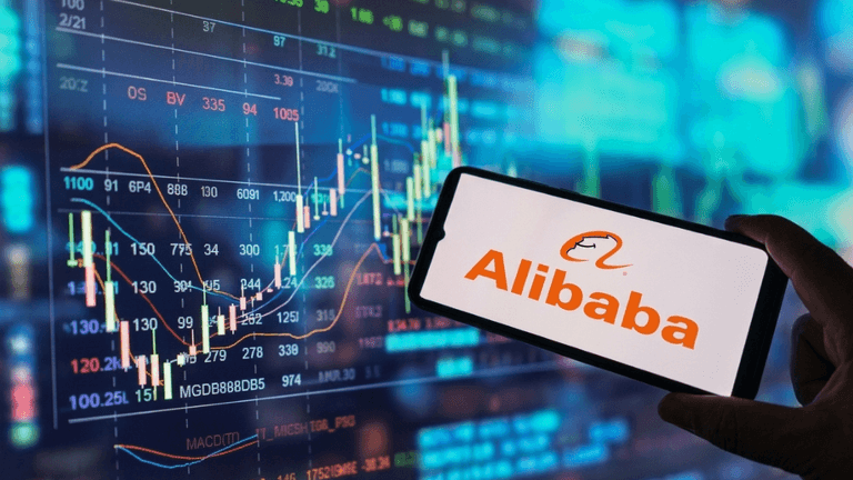Person holding a smartphone displaying Alibaba logo in front of stock market charts