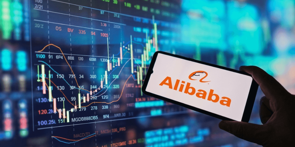 Person holding a smartphone displaying Alibaba logo in front of stock market charts