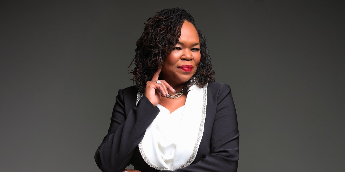 A confident woman in a black blazer with silver trim and a white blouse stands against a grey background, smiling warmly with her hand near her chin.