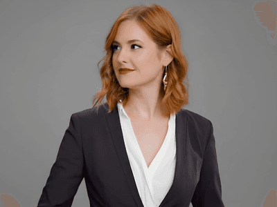 Red-haired woman wearing a dark blazer and white blouse against a gray background.