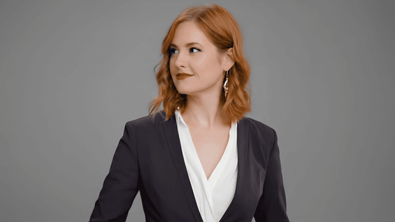 Red-haired woman wearing a dark blazer and white blouse against a gray background.