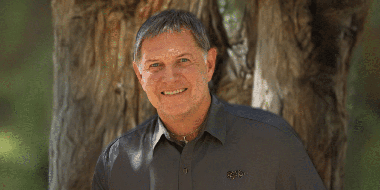 Close-up portrait of a smiling man with short hair, wearing a grey shirt, standing outdoors in front of a tree, showcasing a positive and confident expression.
