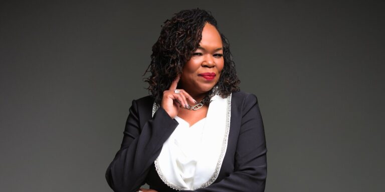 A confident woman in a black blazer with silver trim and a white blouse stands against a grey background, smiling warmly with her hand near her chin.