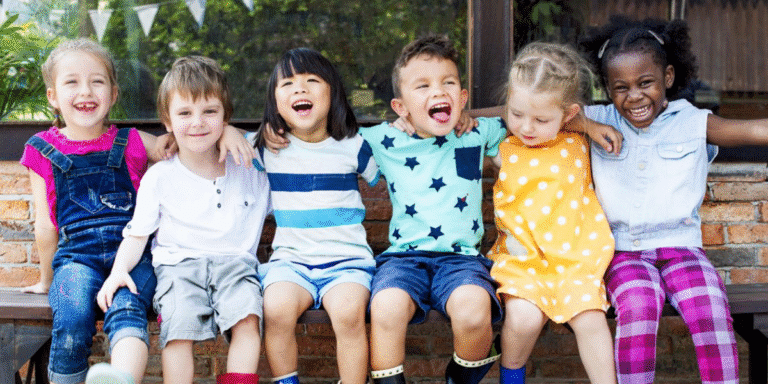 Diverse group of young children sitting together outdoors, smiling and laughing with arms around each other.
