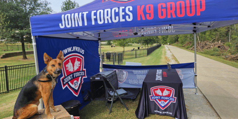 German shepherd at Joint Forces K9 Group booth under blue canopy in park setting.